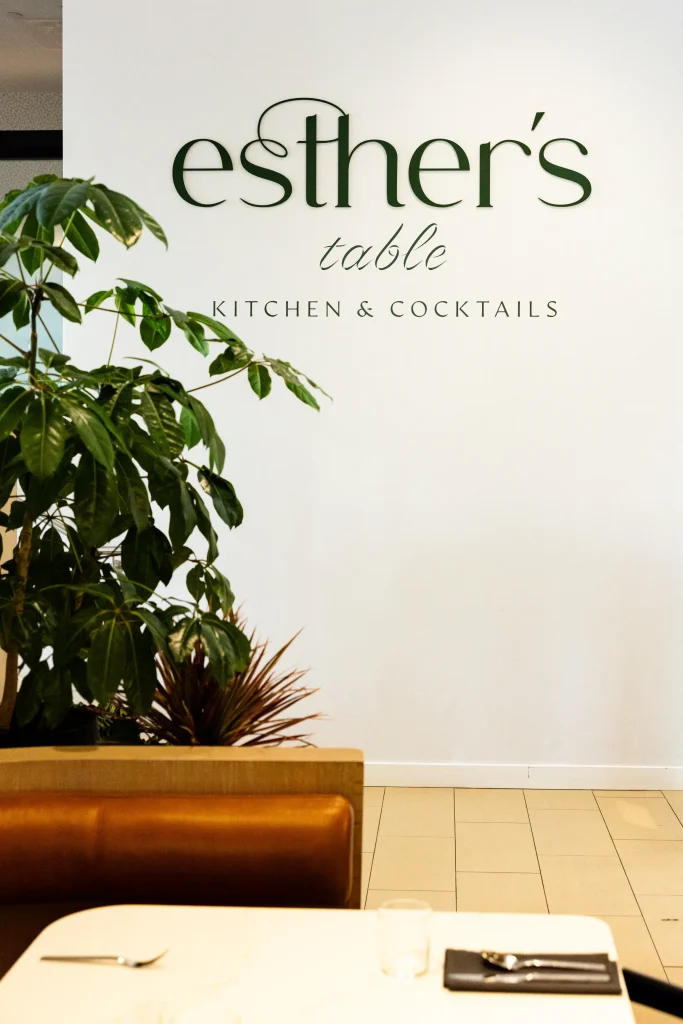 Esther's Table: Bringing a Legendary Minneapolis Hostess Back to the Table 5 Esther's Table signage beside layered live greenery and a cognac leather banquette